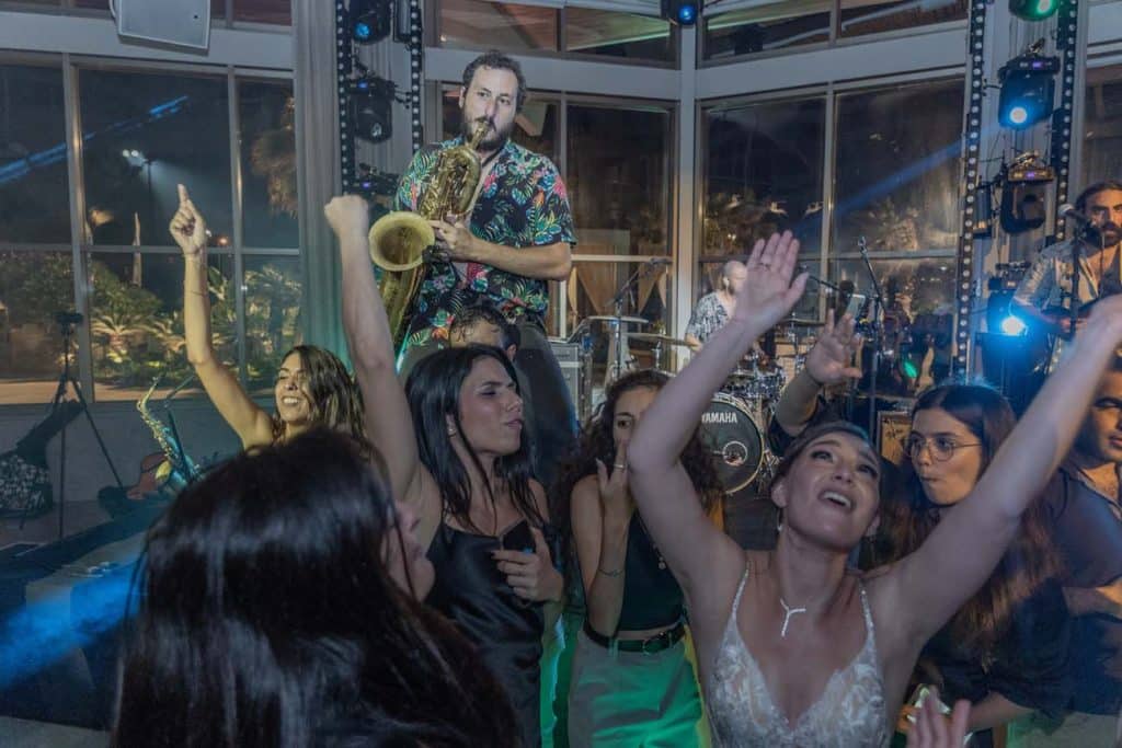 אנשים רוקדים ושמחים בהופעה חיה, עם נגן סקסופון על הבמה ומתופף מאחור.