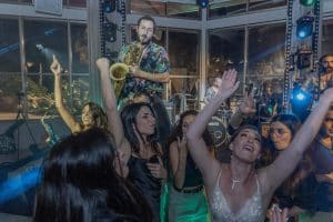 אנשים רוקדים ושמחים בהופעה חיה, עם נגן סקסופון על הבמה ומתופף מאחור.
